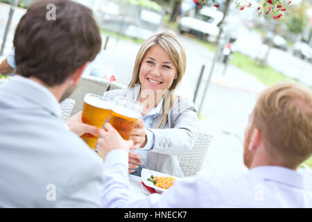 Felice imprenditrice tostare il bicchiere da birra con colleghi di sesso maschile a sidewalk cafe Foto Stock