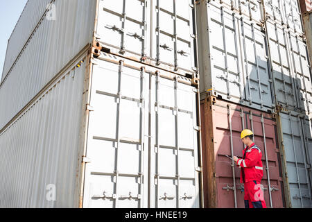 Lavoratore di sesso maschile di ispezionare contenitori di carico durante la scrittura sulla clipboard in cantiere di spedizione Foto Stock