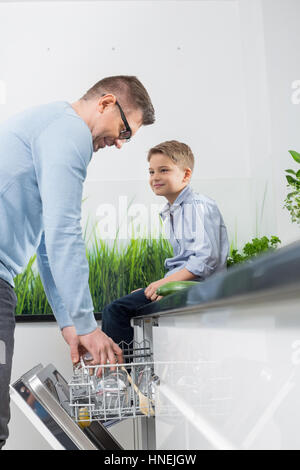 Ragazzo che guarda al padre mettendo il vetro in lavastoviglie in cucina Foto Stock