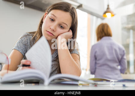 Annoiato ragazza studiare a tavola con la madre in piedi in background Foto Stock