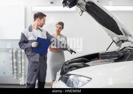 Giovane maschio repairman spiegando auto motore al cliente femmina in automobile repair shop Foto Stock