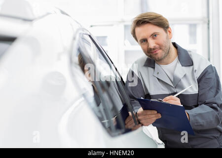 Ritratto di fiducia meccanico di automobile holding clipboard mentre appoggiato su auto della finestra in officina Foto Stock