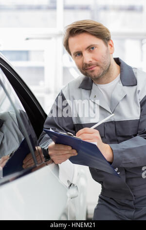 Ritratto di sicuro trattenimento meccanico clipboard mentre appoggiato su auto della finestra in officina Foto Stock