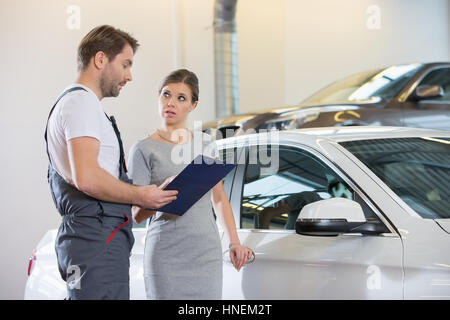 Repairman holding appunti durante la conversazione con il cliente femmina in automobile repair shop Foto Stock