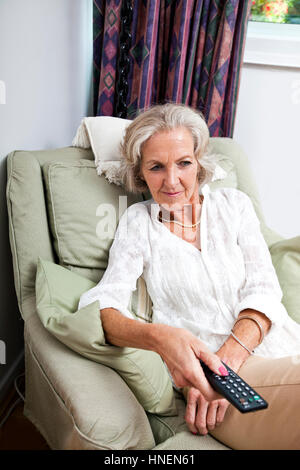 Donna senior di cambiare i canali con telecomando sulla poltrona di casa Foto Stock