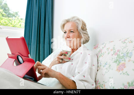 Senior donna utilizzando digitale compressa mentre un caffè sul letto di casa Foto Stock