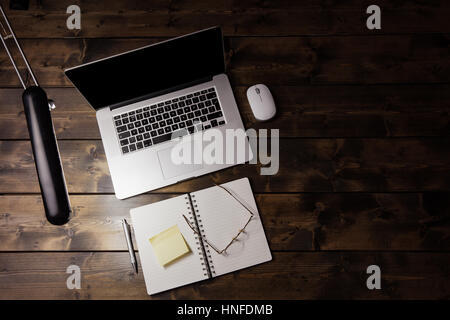 Laptop sulla scrivania in legno visto dal di sopra. Blocco note e gli occhiali da lettura accanto al computer e una scrivania luce illuminando la tabella. Foto Stock