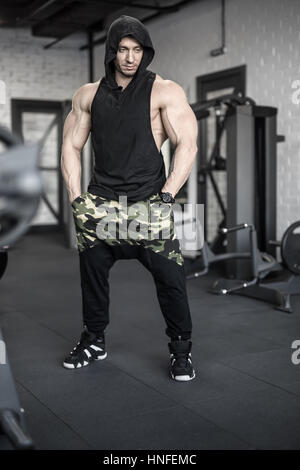 Uomo brutale con il gigante e bicipiti un tatuaggio è in posa nella palestra. Egli indossa una veste nera senza maniche con cappuccio, sneakers e Pantaloni scuri con stampa camouflage. Guy Foto Stock