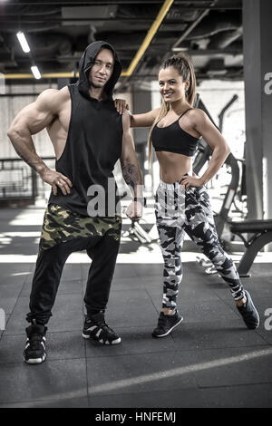 Coppia sorridente stand in palestra. Ragazza indossa multi-pantaloni colorati con un top nero e scarpe da ginnastica, ragazzo indossa pantaloni scuri con un nero con cappuccio e senza maniche Foto Stock