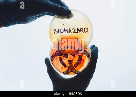 Tenere le mani in capsule di Petri con biohazard parola segno di simbolo. L'acqua contaminata food concept. Pericolose malattie infettive. Medical test di laboratorio di ricerca. Foto Stock