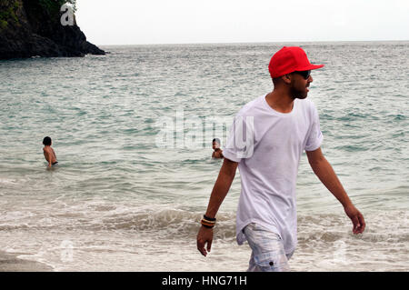 Cool Dude passeggiate lungo la spiaggia sabbiosa in Bali Indonesia indossando Red Baseball Hat, bianco maglietta e pantaloni corti Foto Stock