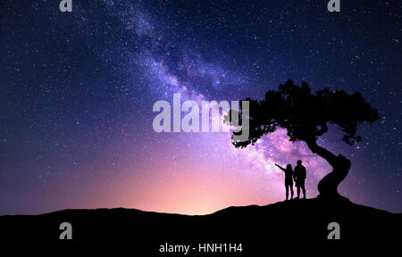 La Via Lattea con persone sotto l'albero sulla collina. Paesaggio con notte cielo stellato e silhouette di felice permanente di un uomo e di una donna che puntare il dito mi Foto Stock