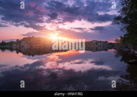 Fiume contro il cielo colorato con le nuvole e rocce al tramonto in estate. Bel paesaggio con il lago, le montagne, la luce del sole e il blu cielo nuvoloso riflessa Foto Stock