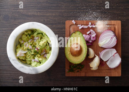 Guacamole Chunky in una coppa di marmo con avocado, l'aglio, il coriandolo, la cipolla e il sale su un tagliere di legno accanto ad esso. Foto Stock