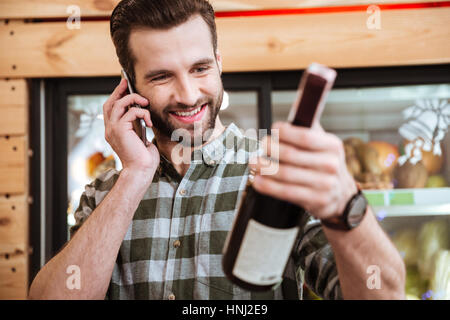 Felice giovane uomo acquistare vino e parlando al cellulare nel negozio di alimentari Foto Stock