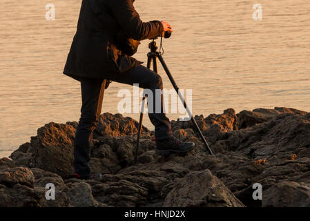 Chiudere fino a un fotografo con un treppiede nella luce del tramonto, fondo di acqua Foto Stock