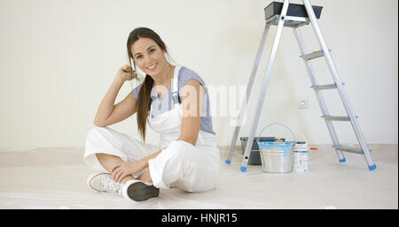 Piuttosto giovane donna rinnovando la sua casa seduto sul pavimento di fronte a una scala e vernice grinning presso la fotocamera Foto Stock