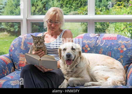 Bionda donna di mezza età è seduto su un divano e la lettura di un libro. Gatto a strisce e Golden Retriever cane sono seduti con il suo insieme. Gli animali sono loo Foto Stock