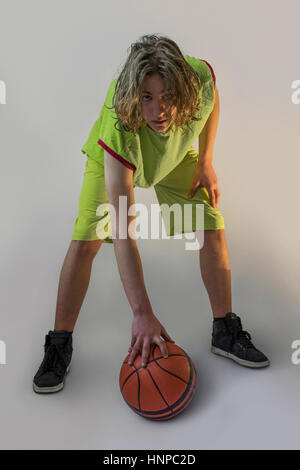 Ragazzo con lunghi capelli biondi che indossa una maglia verde premendo verso il basso una palla da basket e guardando la telecamera Foto Stock