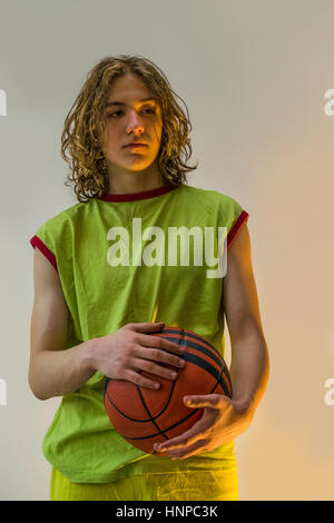 Ragazzo con lunghi capelli biondi che indossa una maglia verde, afferrando una palla da basket e guardando al lato. Foto Stock