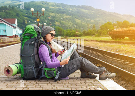 Felice ragazza turistico con zaino siede su una piattaforma in attesa di un treno. Concetto di viaggio. Traveler azienda mappa, in attesa di un treno e piallare per nex Foto Stock