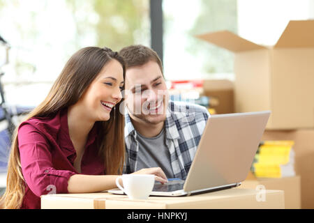 Coppia felice ricerca online con un computer portatile su una scatola di cartone seduta sul pavimento mentre si muove appartamento Foto Stock