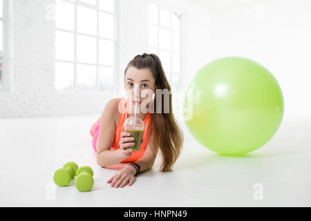 Ritratto di giovane donna sport bere kiwi freschi con pesi e fitness palla sul pavimento nella palestra bianca Foto Stock