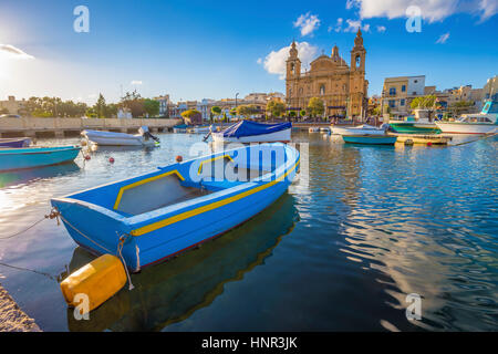 Msida, Malta - Blu tradizionale barca da pesca con il famoso Msida Chiesa Parrocchiale a sfondo su un giorno di estate con cielo blu e nuvole Foto Stock