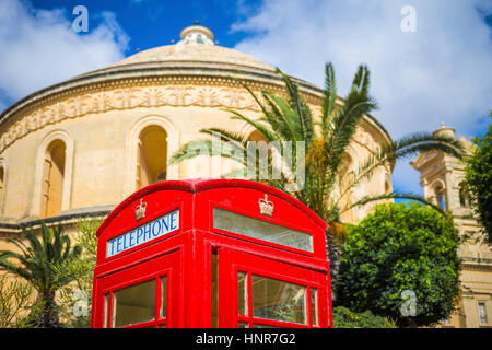 Mosta, Malta - Inglese tradizionale telefono rosso scatola con alberi di palma e il famoso duomo di Mosta a sfondo con cielo blu e nuvole Foto Stock