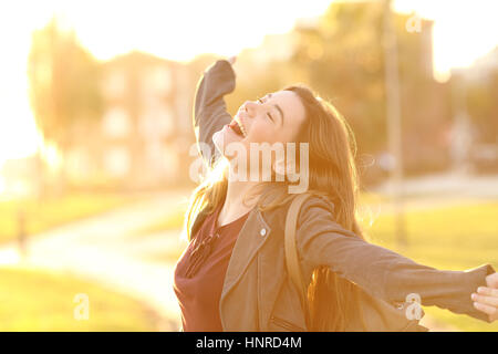 Ritratto di un adolescente eccitato ragazza i bracci di sollevamento e ridere in strada al tramonto con una luce calda in background Foto Stock