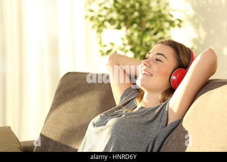 Un rilassato riposo teen e ascolto di musica con cuffie wireless seduto su un divano nel soggiorno di casa con una luce calda Foto Stock