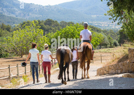 Vista posteriore della femmina groom leader cavaliere a cavallo lungo il paddock a scuderie rurale Foto Stock