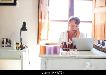 Donna che parla sullo smartphone mentre utilizzando laptop nel sapone artigianale ufficio di officina Foto Stock