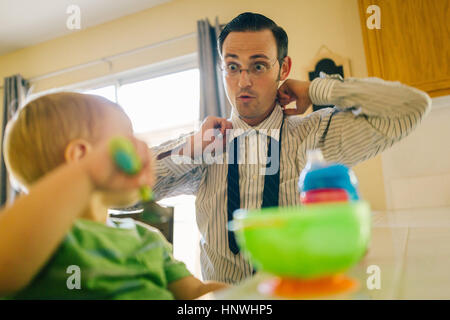 Padre e figlio giovane in cucina, padre mettendo sul Tie collo mentre figlio mangia la prima colazione Foto Stock
