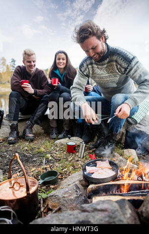 L'uomo la cottura di cibo sul fuoco con gli amici in background Foto Stock