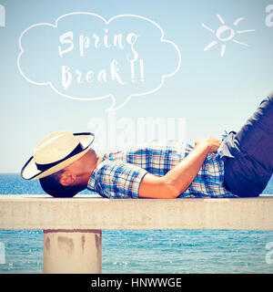 Un giovane uomo caucasico con un cappello di paglia sul suo volto disteso in un banco di strada vicino al mare e testo spring break in un palloncino di parlato Foto Stock