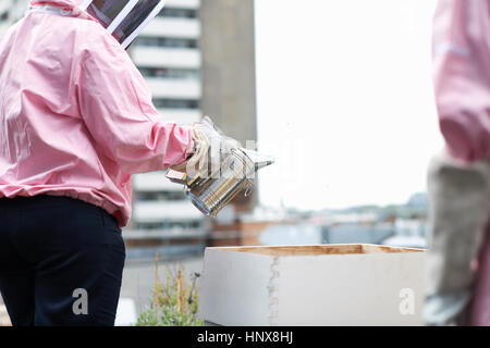 Apicoltore in piedi accanto a hive, tenendo bee fumatore, metà sezione Foto Stock