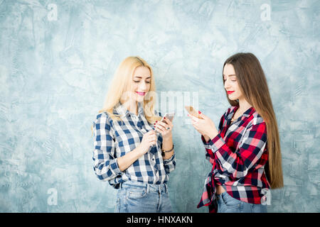 Amici di sesso femminile in camicie a scacchi utilizzando i telefoni intelligenti sulla parete blu sullo sfondo Foto Stock