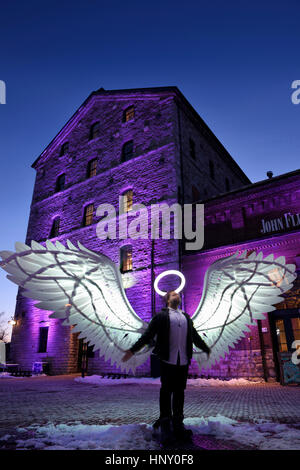 Uomo con leggero alone e angelo ali in corrispondenza storico mulino in pietra edificio a Distillery District Toronto Light Festival in inverno al tramonto Foto Stock