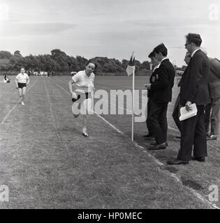 Anni sessanta, storico, una determinata scuola girl circa a tagliare il traguardo in una 100 yard in esecuzione fuori gara su una pista in erba. Foto Stock