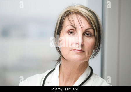 Frontale di testa e spalle ritratto di una donna di mezza età in camice bianco con uno stetoscopio prima di una finestra in posizione eretta e seriamente guardando nella Foto Stock