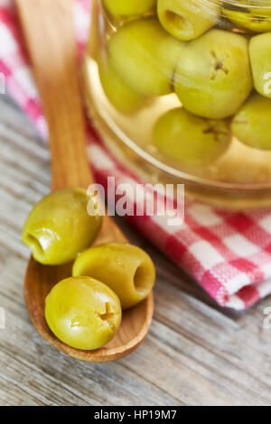 Decapati olive verdi in cucchiaio di legno accanto a un vaso di olive da conserva Foto Stock