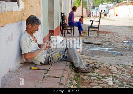 Un cubano uomo seduto sul marciapiede in ombra della sua casa lavorando su alcune di corda in Trinidad, Cuba Foto Stock