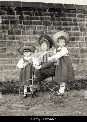 Ritratto di tre bambini in stile vittoriano su un triciclo di fronte a un muro di mattoni. Fotografia scattata nel 1900, restaurata da una scansione ad alta risoluzione prese dal negativo originale. Foto Stock