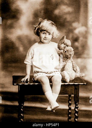 Formale di Edwardian ritratto di un piccolo bambino seduto su un tavolo basso, indossando un abito di maglia e coccole il suo giocattolo coniglio. Dietro la ragazza ci sono tre passi e ciò che appare essere un fotografo studio del foglio di sfondo. Fotografia scattata nel 1910, restaurata da una scansione ad alta risoluzione prese dall'originale Edwardian negativo. Foto Stock