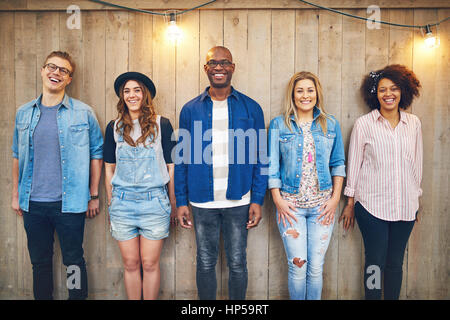 Cinque giovani uomini e donne in abbigliamento casual permanente ancora in una fila in legno parete non verniciata, sorridente e guardando la fotocamera Foto Stock
