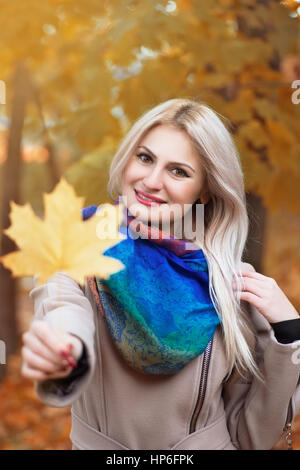 Ragazza con autunno orange Foglia di acero. Ritratto di giovane e bella donna bionda con Foglia di acero in mano di trascorrere del tempo in autunno park con il sole Foto Stock