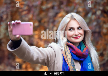 Bella bionda e giovane donna caucasica tenendo selfie con lo smartphone all'aperto nel parco in autunno. Selfie autunno. In autunno, persone, tecnologia, stile di vita Foto Stock