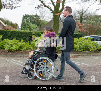 L'uomo spinge una donna su una sedia a rotelle in un posto di parcheggio in un villaggio olandese Foto Stock