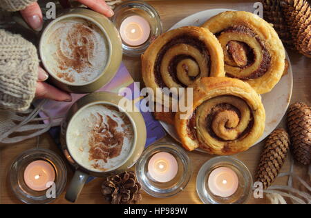 Una donna raggiunge per una cremosa mocha (caffè e cioccolato) servita con ciambelle alla cannella) servita a lume di candela in un accogliente English home in inverno Foto Stock